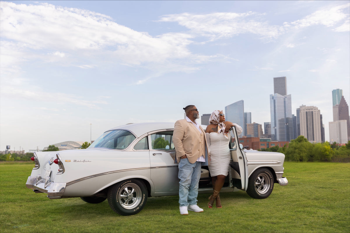 Rear three-quarter view of a white 1956 Chevrolet Bel Air with a couple standing beside the open passenger door and Houston buildings behind them.